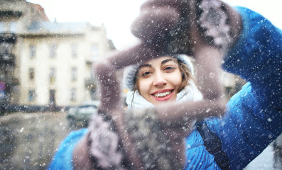 portrait of a young smiling attractive woman in warm winter clothes at sowy day on the street background. woman shows a frame from hands like photo. Photo Frame Hands Made By A Hipster young girl © vitaliymateha