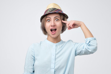 Surprised smiling young woman in summer hat, twisting her index finger at temple
