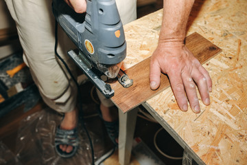 Worker is holding in hands power jigsaw and cutting laminate flooring in apartment. Maintenance repair works renovation in the flat with scroll saw. Restoration of wooden parquet floor planks indoors.