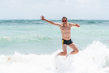 Young happy man jumping up with arms raised waving in red swimsuit bikini bathing suit and splash crashing wave in Miami Beach Florida green water drops