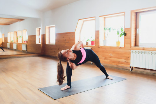 Rear view of young female yogi in sports bra and leggings doing bound extended side angle pose, baddha utthita parsvakonasana, while practicing yoga indoors.