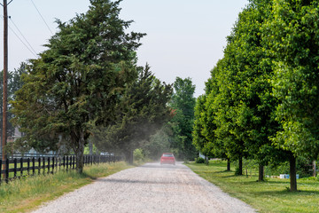 Summer road during day in Virginia rural farm countryside with red car and dust on dirt road, fence and trees row lined