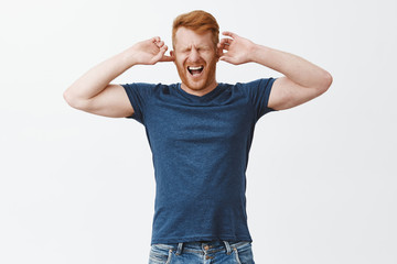 Fototapeta premium Displeased redhead guy feeling pain in head cause of noisy annoying sound, covering ears with fingers, shouting from painful feeling, closing eyes and yelling with opened mouth over grey wall