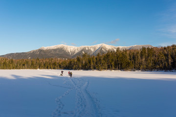 a group of hikers with a dog walk across a snowy frozen lake on there way to the snop capped White Mountains