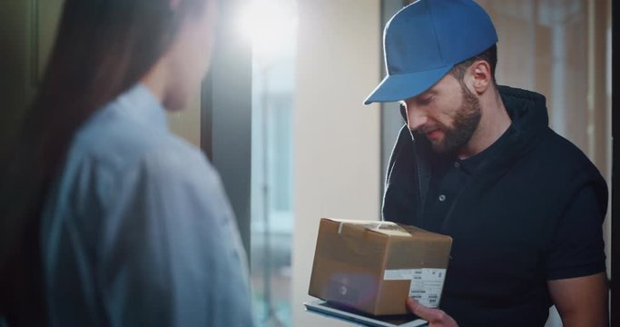 Happy young woman receiving a parcel from mailman delivered in her home. Shot with RED camera i 8K. Concept of courier, delivery, e-commerce, online shopping