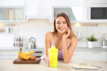 Young woman holding bottle of protein shake at table with ingredients in kitchen