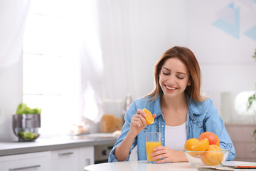 Happy young woman squeezing orange juice into glass at table in kitchen, space for text. Healthy diet