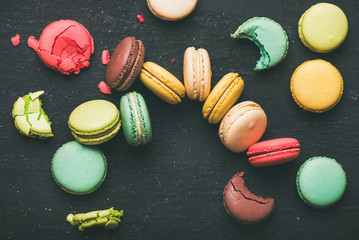Flat-lay of sweet colorful French macaroon cookies variety with sugar powder over black background, top view, horizontal composition