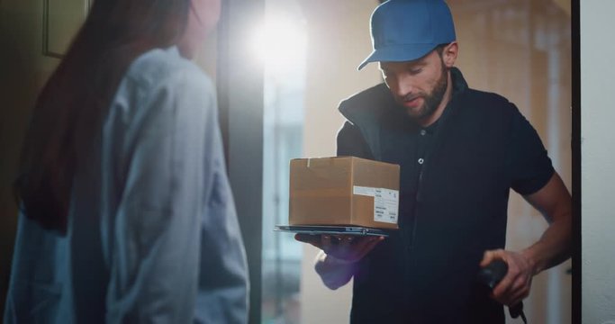 Happy young woman receiving using smartphone for digital signing a parcel from mailman delivered in her home. Shot with RED camera i 8K. Concept of courier, delivery, e-commerce, online shopping