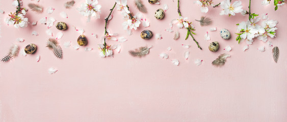 Easter holiday background. Flat-lay of tender Spring almond blossom flowers on branches, feathers, quail eggs over light pink background, top view, copy space, wide composition. Greeting card concept