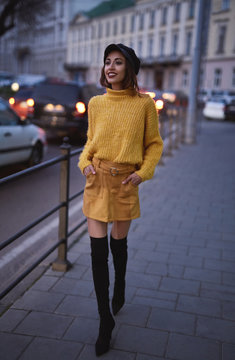 vertical portrait of a beautiful fashionable stylish woman in bright yellow sweater and skirt, black high heel boots and cap walking and posing outdoors at evening, street style shooting