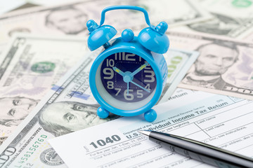 Time or deadline for yearly tax submission concept, miniature small retro alarm clock with pen on 1040 US individual income tax filling form on pile of US dollar banknotes money