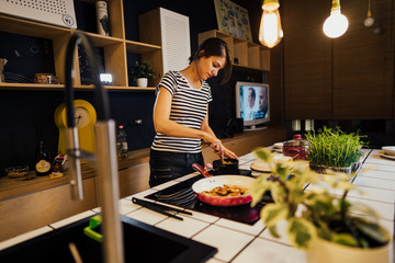 Young house wife cooking a healthy meal in home kitchen.Making dinner on kitchen island standing by induction hob.Preparing fresh vegetables,enjoying spice aromas.Passion for cooking.Paleo diet