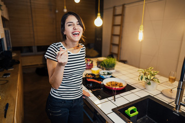 Young woman tasting a healthy meal in home kitchen.Making dinner on kitchen island standing by...