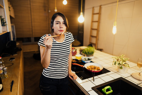 Young woman tasting a healthy meal in home kitchen.Making dinner on kitchen island standing by induction hob.Preparing fresh vegetables,enjoying spice aromas.Eating in.Passion for cooking.Keto diet