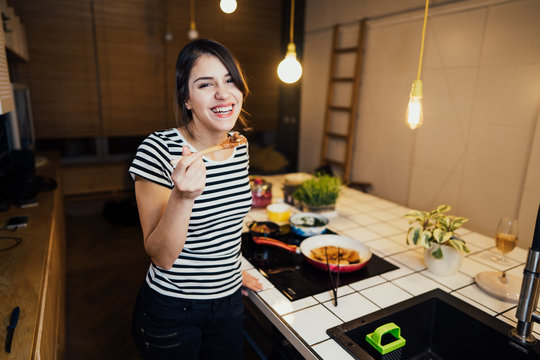 Young woman tasting a healthy meal in home kitchen.Making dinner on kitchen island standing by induction hob.Preparing fresh vegetables,enjoying spice aromas.Eating in.Passion for cooking.Dieting 