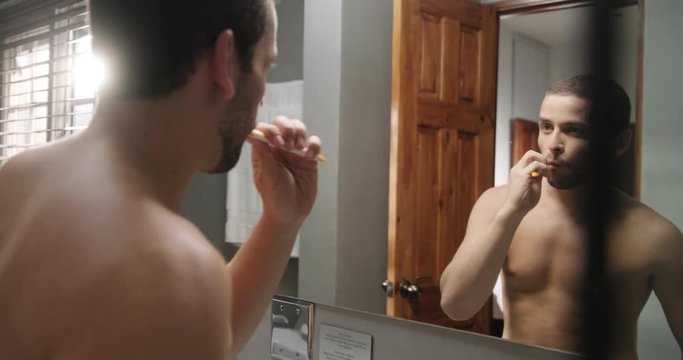 Young homosexual hispanic people and male beauty. Beautiful latino person grooming in bathroom in hotel room for morning routine, body care. White gay metrosexual man brushing teeth in front of mirror