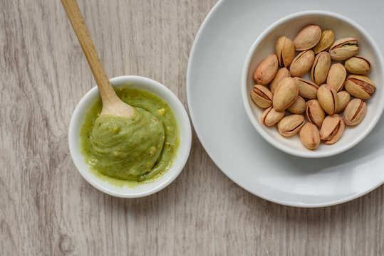 Still life of pistachio paste and unpeeled salted pistachios in a white plates, wooden spoon for pasta on a light background. Components for cooking.