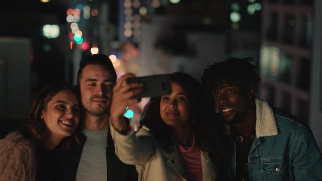 young multi ethnic friends on rooftop at night posing for group photo celebrating friendship reunion young woman using smartphone sharing weekend gathering to social media