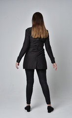  full length portrait of a brunette girl wearing long black coat, standing pose with back to the camera, on grey studio background.