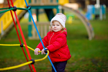 Cute toddler girl having fun on playground. Happy healthy little child climbing, swinging and sliding on different equipment. On cold day in colorful clothes. Active outdoors game for children