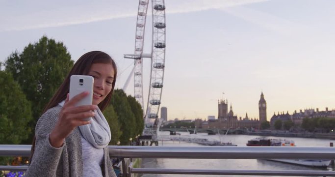 London travel woman taking selfie with mobile phone camera. Young Asian tourist or casual business woman smiling at camera with Thames river, Big Ben and Westminster bridge and london eye background.