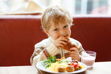 Cute healthy kid boy eating croissant and drinking strawberry milkshake in cafe. Happy child having breakfast with parents or at nursery. Vegetables, eggs as healthy food for children.