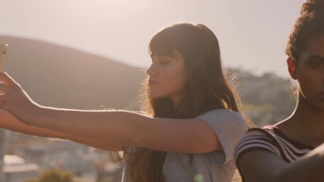 beautiful young women friends taking selfies using smartphone enjoying hanging out on rooftop at sunset ready for weekend party