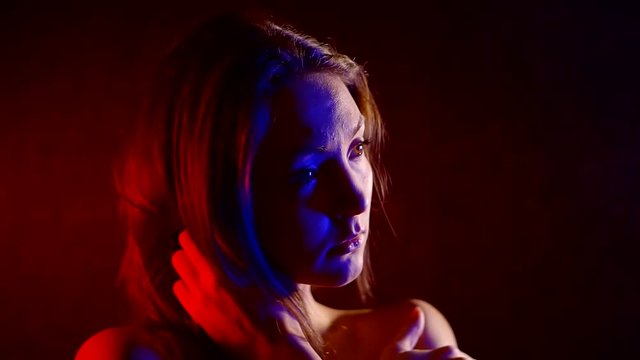 sad and anxious young woman is hugging herself in dark room, red and blue lights are lighting on her
