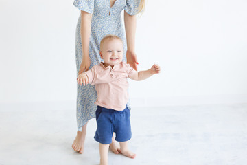 First steps - young mother in a blue dress is holding the hands of a small charming son in a T-shirt and shorts on a white background. Place for text
