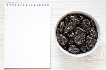 Organic dry prunes and blank notepad on a white wooden surface, top view. Overhead, flat lay, from above. Space for text.