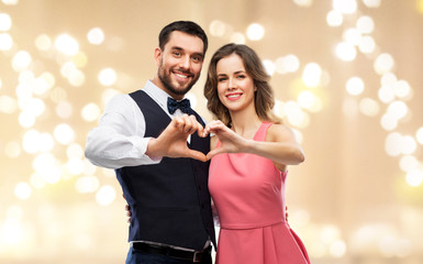 valentines day, love and people concept - happy couple in party clothes making hand heart gesture over beige background with festive lights