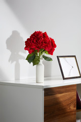 Side shot of an interior design. There are a photoframe and a vase with red hydrangea bouquet on table. The floral composition is isolated in the middle on white background, making a shadow on wall.