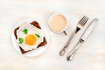 An egg cooked sunny side up on a toast with coffee, shot from above with a fork and knife, and a...