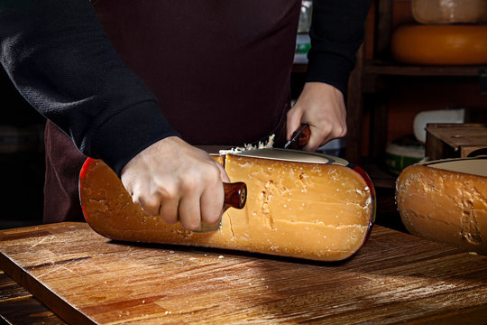Young worker man cuts half of wheel gauda cheese in shop. Wooden background, knife for cheese. Close up view. Copy space for text, logo or brand. Work Concept.
