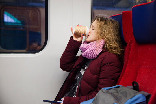 A young girl with curly hair sits in a train car and and drinking coffee from a paper cup
