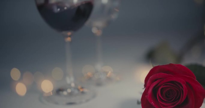 Close-up dolly shot. A single red rose. Two glasses of are beng filled with wine. One glass is lifted by a male hand. Bokeh background.