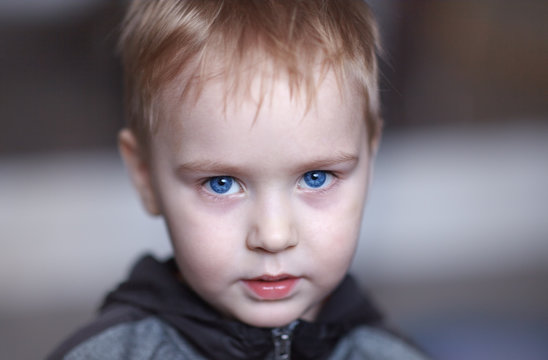 Close up portrait of cute caucasian baby boy with very serious face expression. Bright blue eyes, fair hair. Strong emotions. Indoors, copy space.