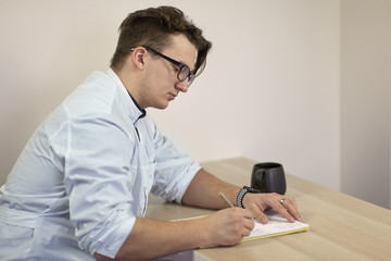 Young caucasian male doctor in a white outfit writes something sitting on the table with cup of tea or coffee. Serious face expression, glasses, finger ring. Indoors, copy space.