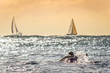 Male surfer paddling out at sunset in Hawaii with sailboats in the background