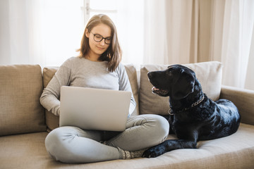 Teenage girl with a dog sitting on a sofa indoors, working on a laptop.