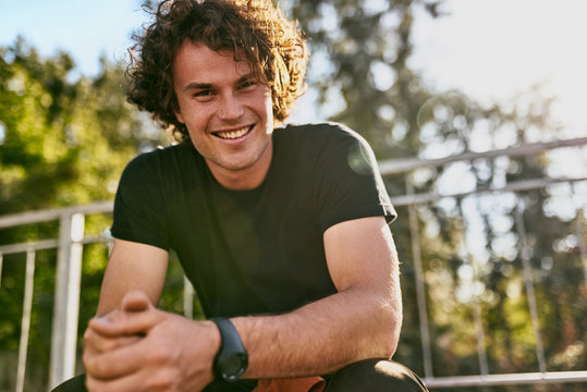 Portrait Of Smiling Cheerful Young Man Wears Black T-shirt And Wristwatch On The City Street. Happy Male With Curly Hair Posing For Advertisement With Copy Space Outdoor. People Concept