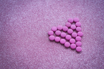 Obraz na płótnie Canvas pink pills laid out in shape of a heart on glitter pink background. coloured drugs. concept - heart disease, heart disorders and drugs, cardiology, valentines, love,