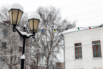 Street lamp under the snow and a garland of multicolored light bulbs tied to it against the background of white buildings, birches and white sky. Winter, Veliky Ustyug, Russia