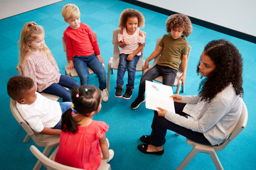 Young female school teacher reading a book to infant school children, sitting on chairs in a circle...
