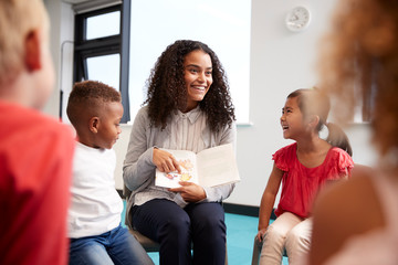 Young female teacher showing a picture in a book to children in an infant school class sitting on...