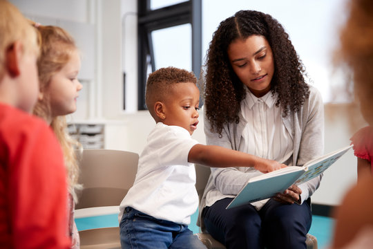 Infant School Boy Pointing In A Book Held By The Female Teacher, Sitting With Kids On Chairs In The Classroom, Close Up