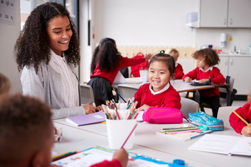 Female teacher and Chinese schoolgirl sitting at a table in an infant school class smiling to other...