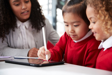 Close up of female infant school teacher sitting at a desk in a classroom helping two schoolgirls wearing school uniforms using a tablet computer and stylus, looking at screen and smiling