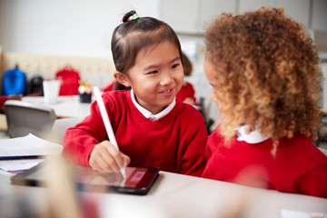 Close up of two kindergarten schoolgirls wearing school uniforms, sitting at a desk in a classroom using a tablet computer and stylus, looking at each other smiling